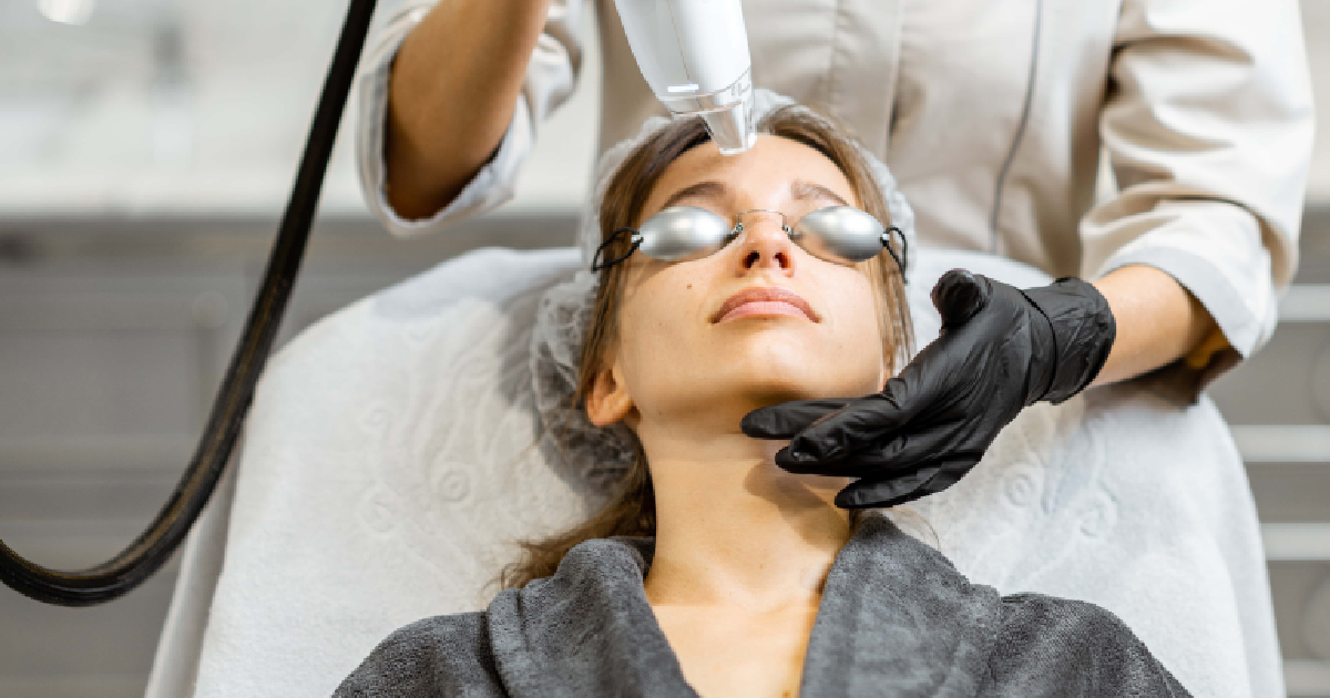 Woman receiving IPL Photofacial in Long Beach, CA, wearing protective goggles while a professional uses a handheld light-based device on her face.